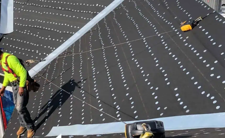 Worker in safety harness installing black waterproof sheeting on commercial roof