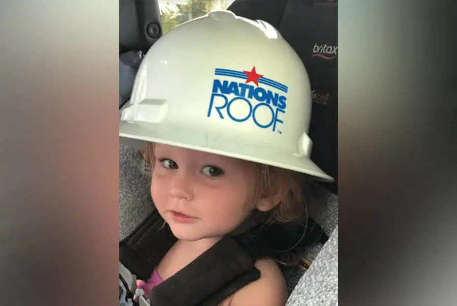 Young child wearing "Nation's Roof" hard hat, smiling in car seat