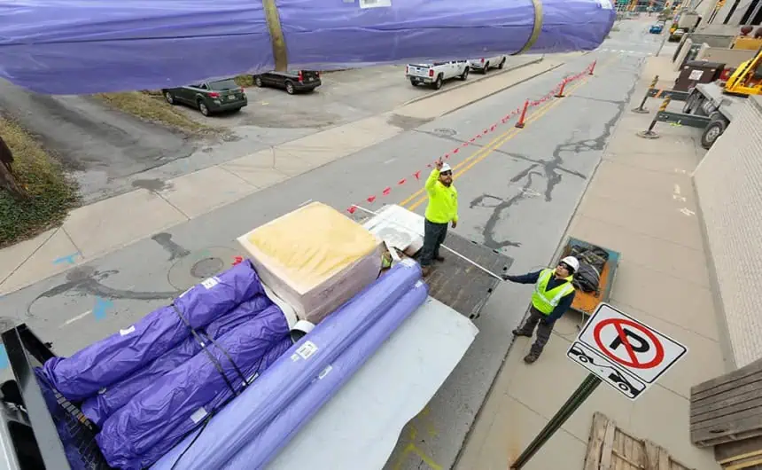 Crane lifting materials for commercial roof installation, guided by workers in safety vests.