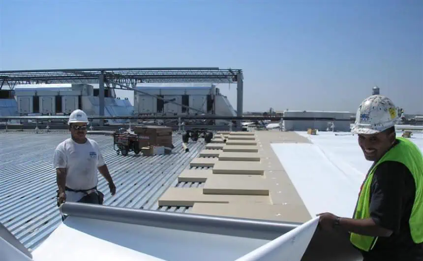 Workers unrolling TPO membrane on flat commercial roof under clear skies.