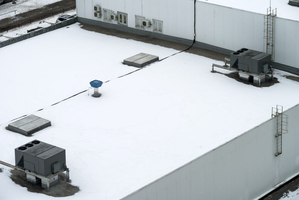 Multi-level commercial flat roof with air handlers and skylights covered in snow.