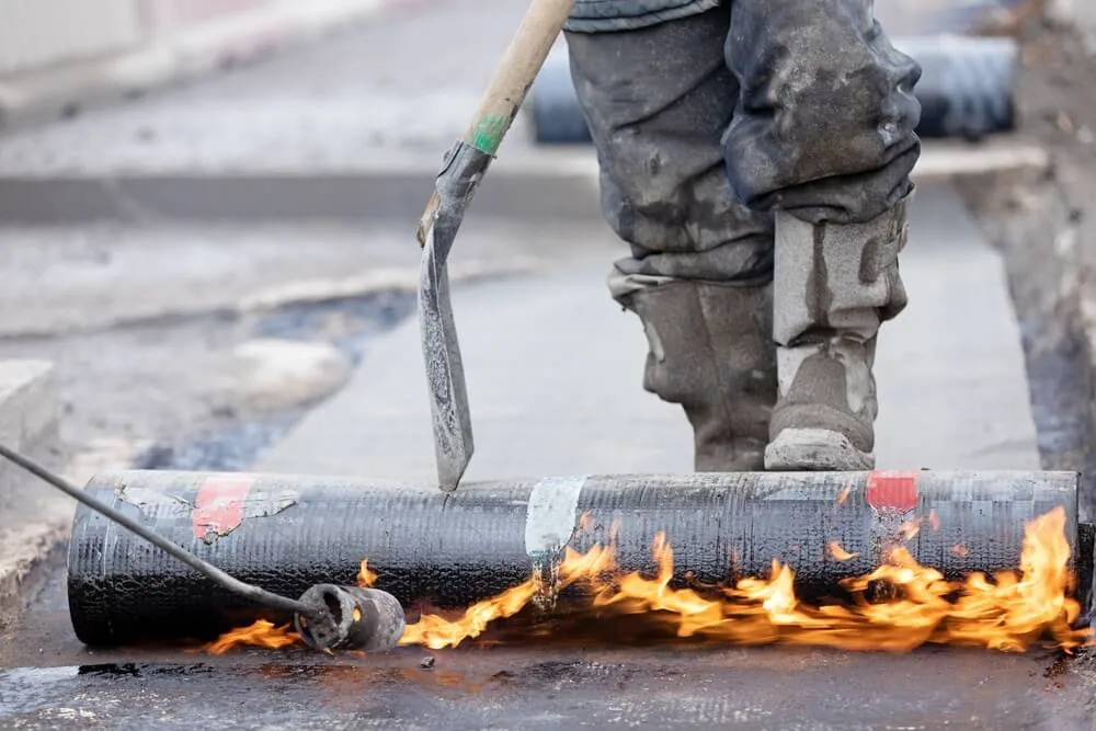 Two works apply a cap sheet with torches on a two-ply roof.